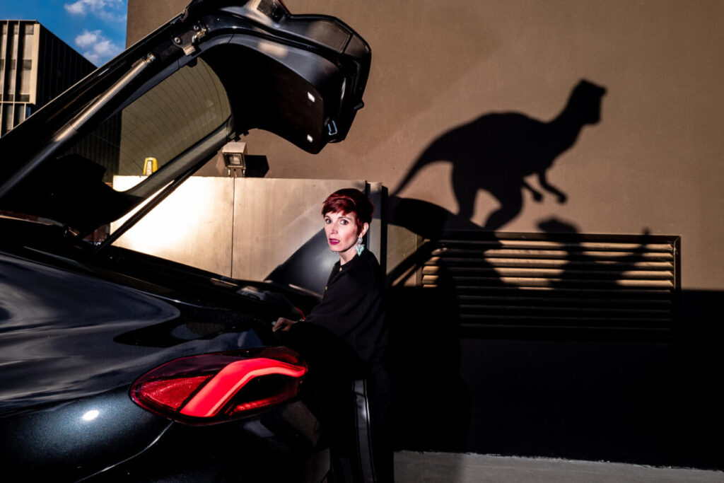 Jurassic Street, donna dietro a una macchina con cofano aperto di alto, sullo sfondo ombre di velociraptor - foto di Massimiliano Faralli