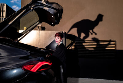 Jurassic Street, donna dietro a una macchina con cofano aperto di alto, sullo sfondo ombre di velociraptor - foto di Massimiliano Faralli