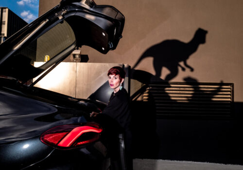 Jurassic Street, donna dietro a una macchina con cofano aperto di alto, sullo sfondo ombre di velociraptor - foto di Massimiliano Faralli